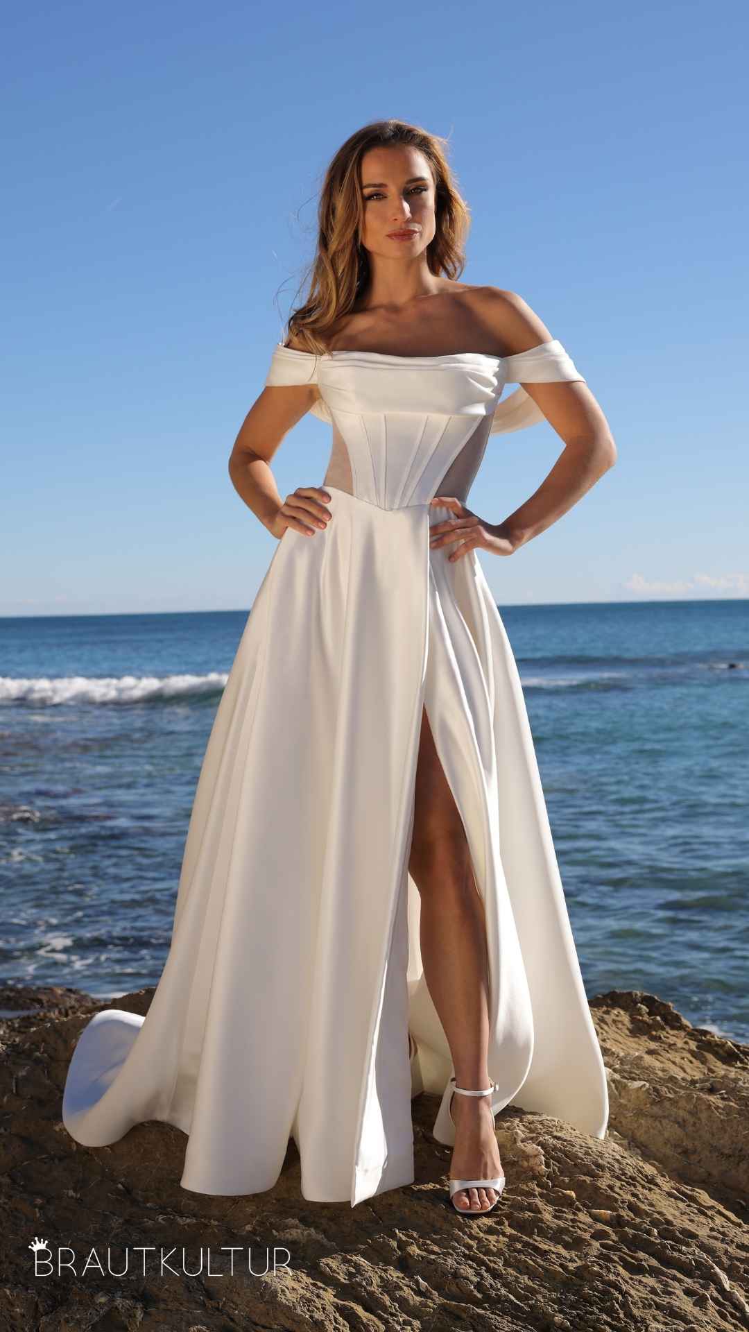 Designer Brautmode ganz in der Nähe von Köln. Dieses glatte Brautkleid aus hochwertigem Satin ist schlicht, aber auch sehr elegant und wunderschön. Das Brautkleid von Azuree hat Carmen Träger und einen Schlitz im Rock.